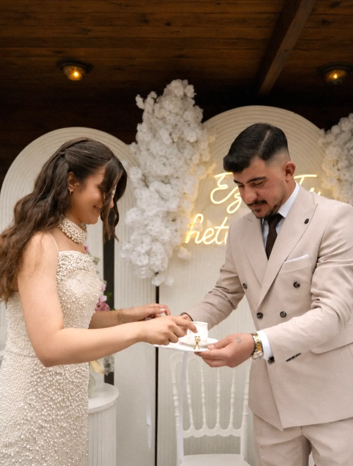 At the engagement the couple pass a small tray between them in front of a white flower wall