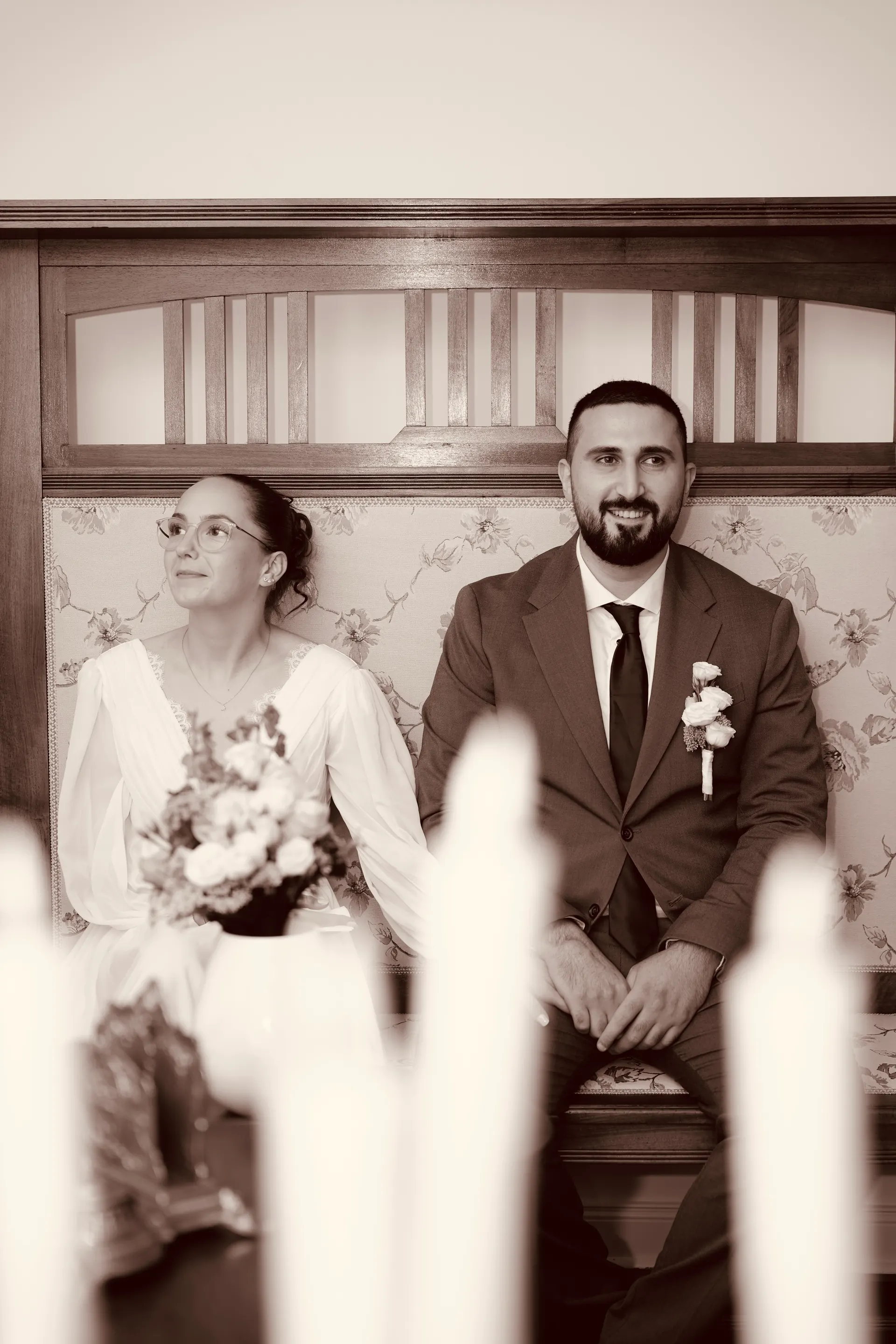 Warm-toned black and white: bride and groom sit on a bench in the registry office, candles blurred in front